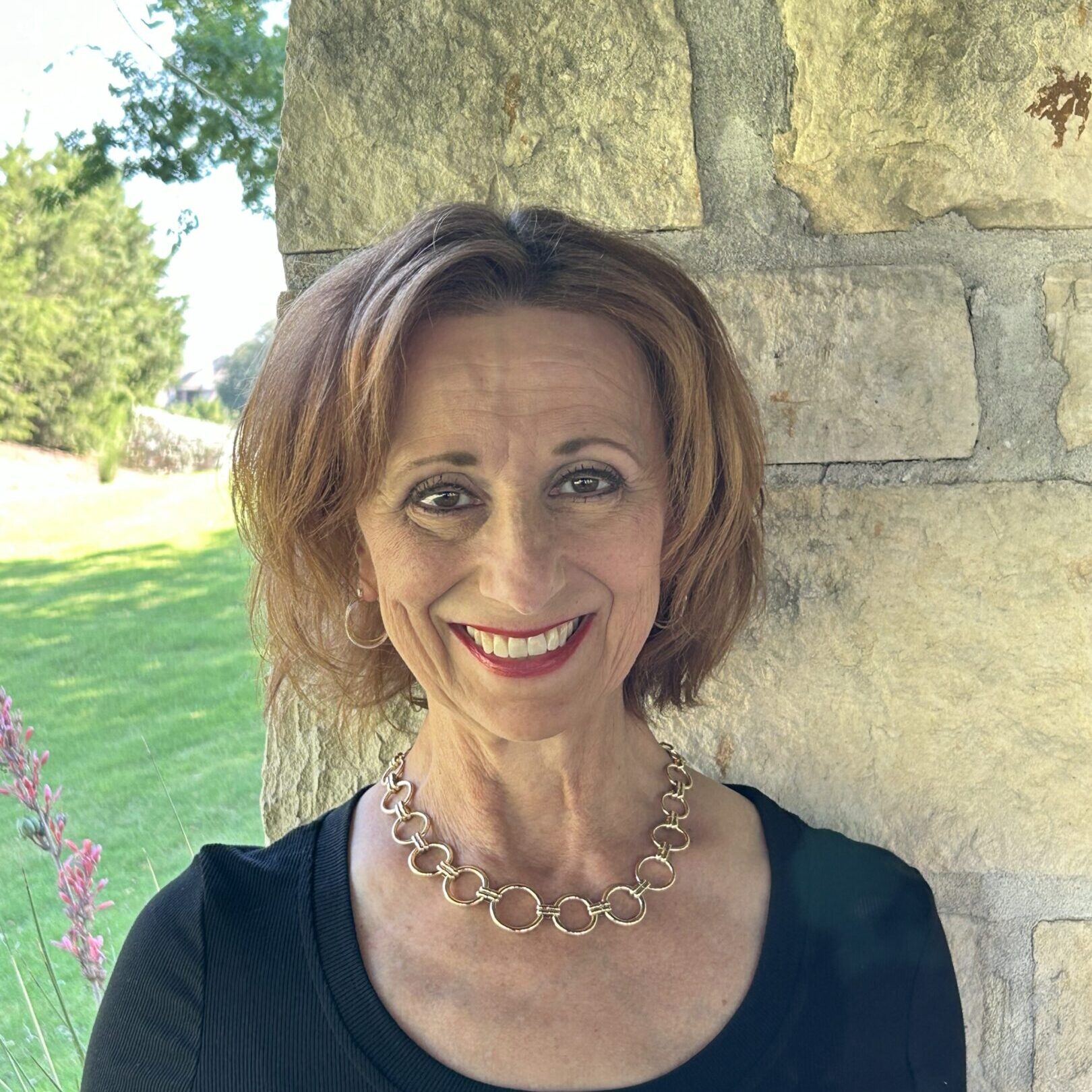 Smiling woman with short hair posing outdoors by a stone wall.